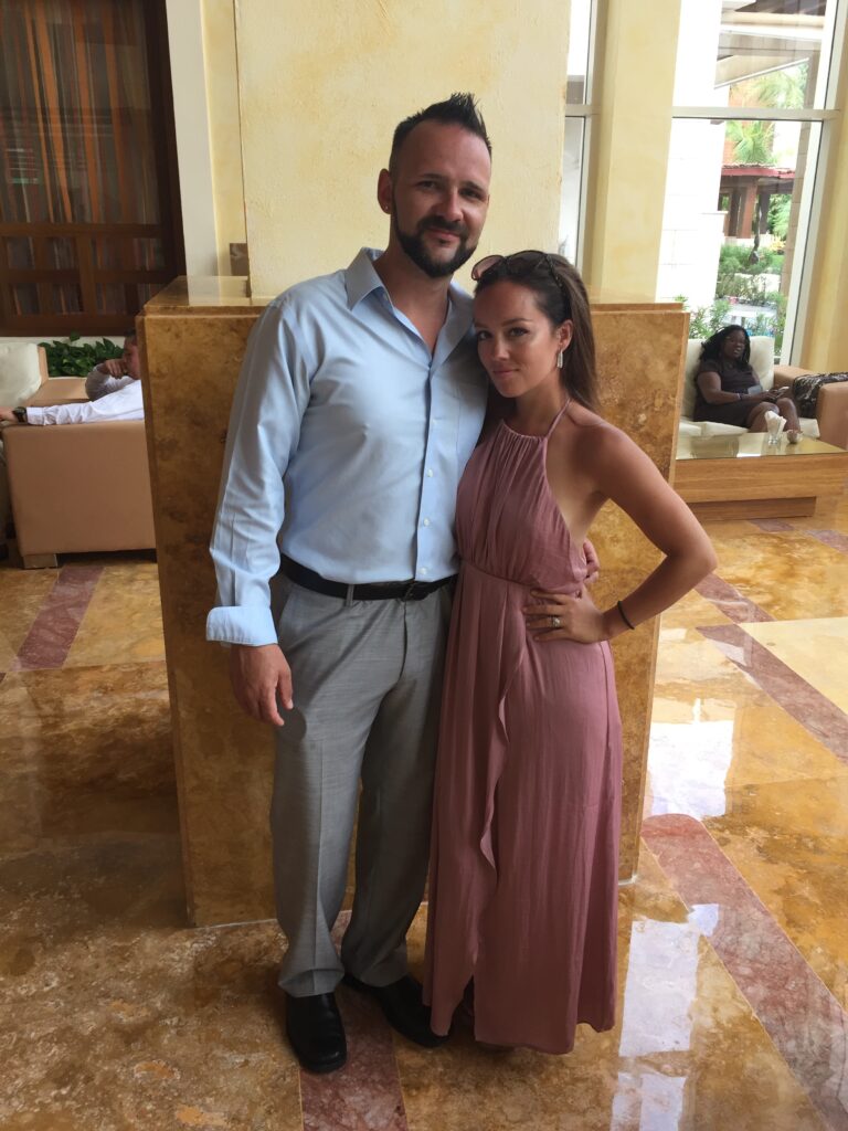 A man and a woman smile at the camera in the lobby of a tropical resort.