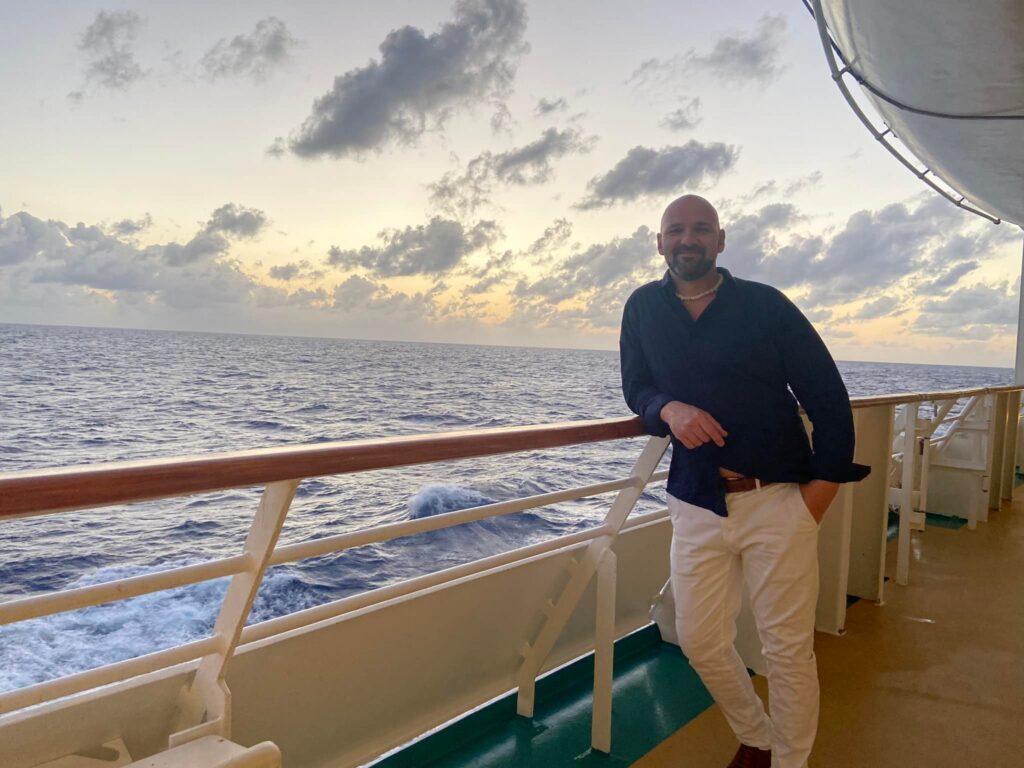 A man leans on the railing of a cruise ship at sunset.