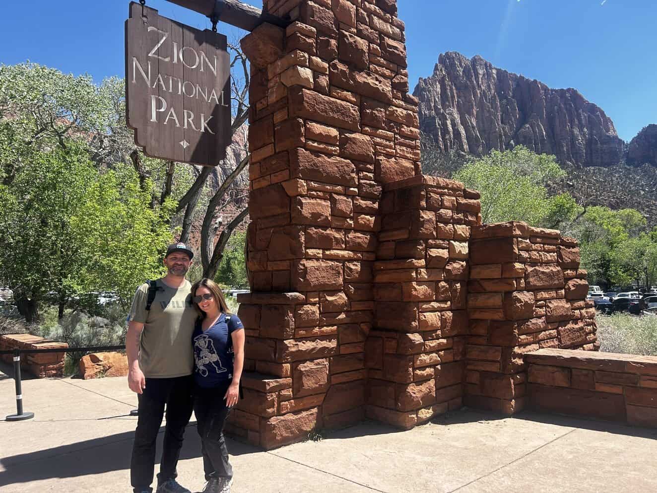 a man and woman under a big brown Zion National Park sign