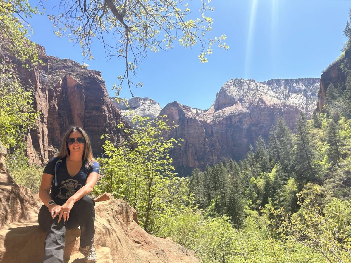 A woman sitting on a rock in Zion National Park with lush green trees and blue sky in the background.