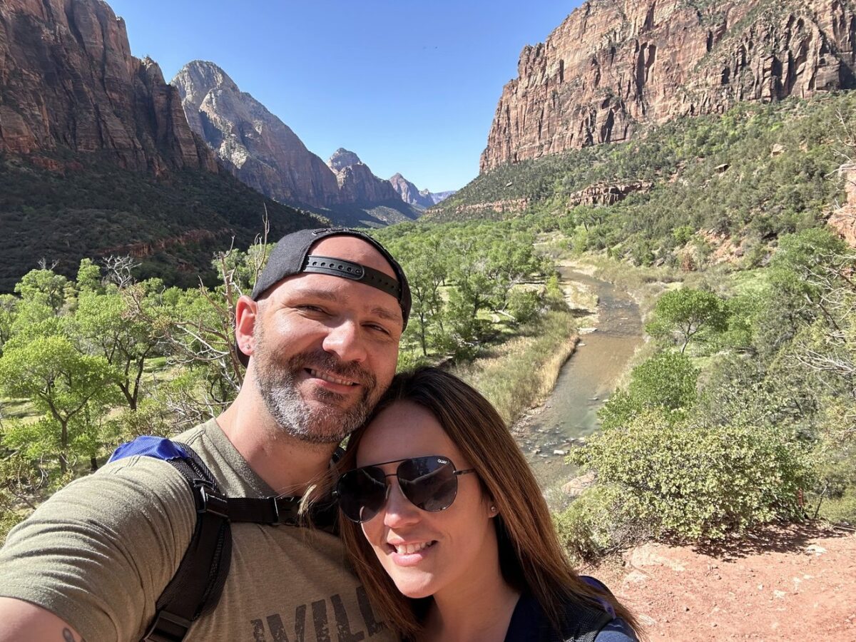 A man and woman in the foreground with a beautiful background in Zion National Park of cliffs, lush green trees, and blue sky.